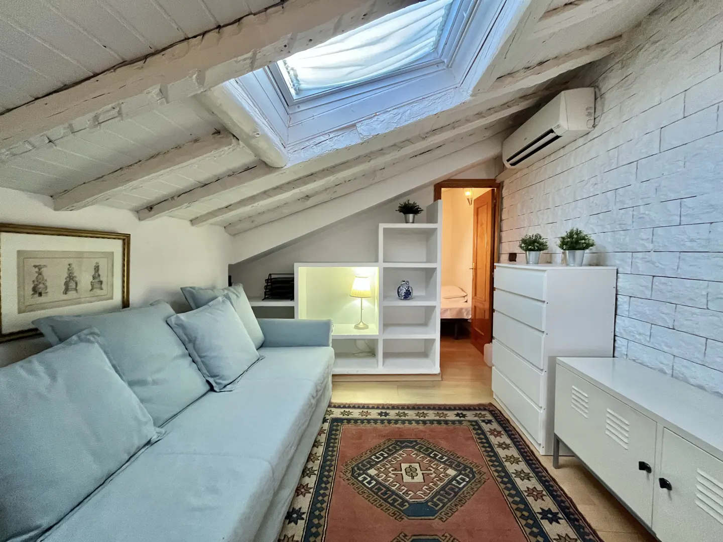 Attic living room with a light blue sofa, white brick wall, skylight, patterned rug, and built-in shelving.