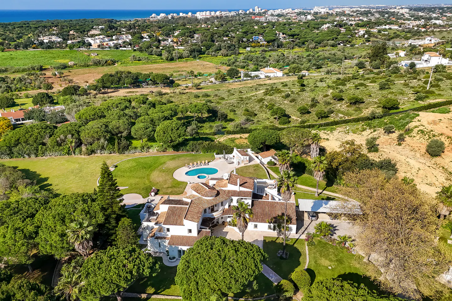Aerial view of a large white villa with a red tile roof, surrounded by green trees and a blue pool. The ocean and city skyline are visible in the background.