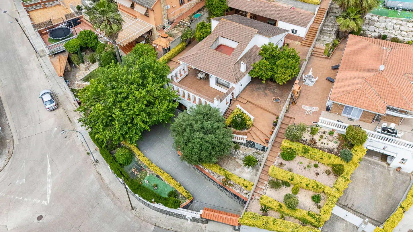 Aerial view of a white villa with a brown tiled roof, large trees, a paved driveway, and terraced gardens.
