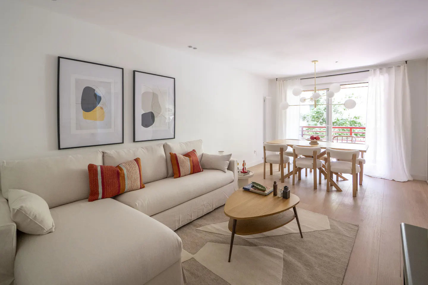 Bright living room with a white sofa, orange pillows, and abstract art. A dining table and chairs are visible in the background.