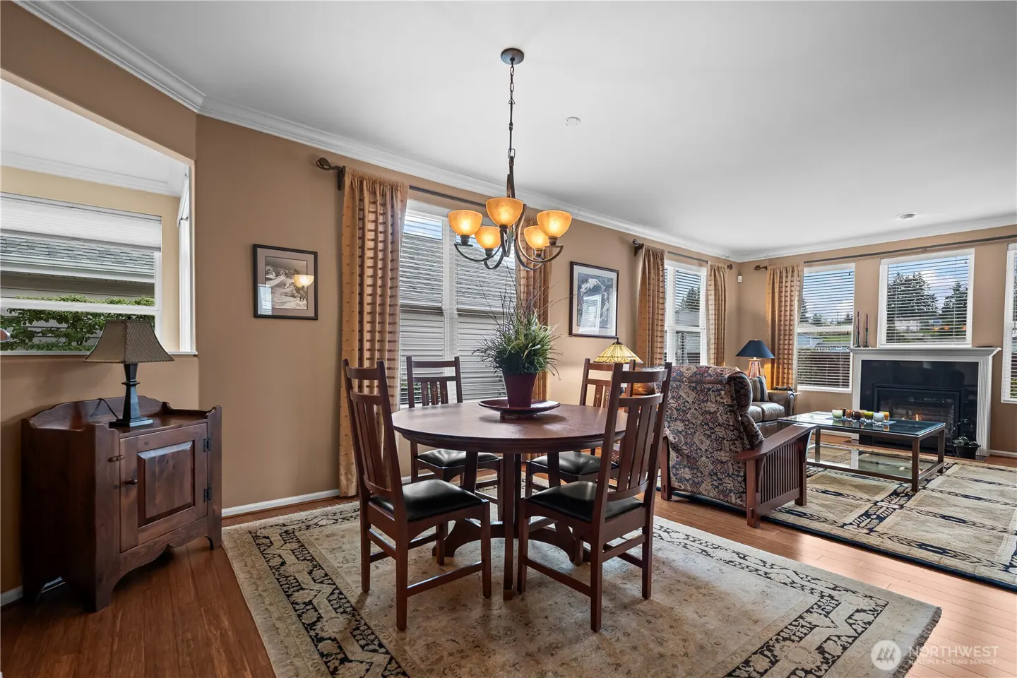 A dining room with a round wooden table, chairs, and a chandelier. The room has tan walls, hardwood floors, and a patterned rug. A living room with a fireplace is visible in the background.