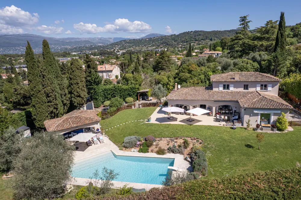Aerial view of a luxury home with a pool, patio, and lush green lawn, set against a backdrop of rolling hills and a clear blue sky.