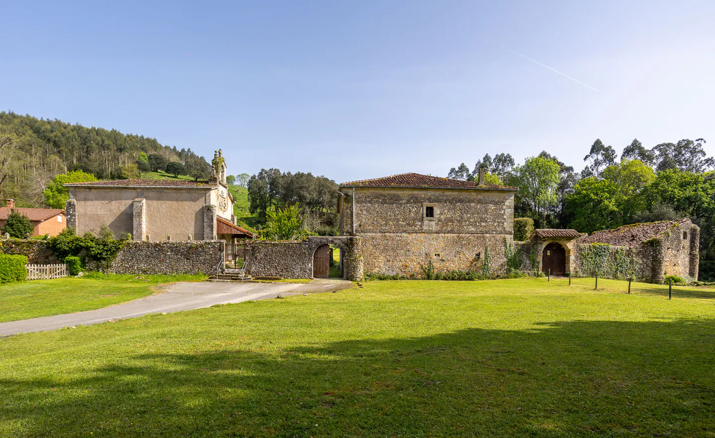 Exterior view of a stone building with a red tile roof, a church, and a green lawn on a sunny day.