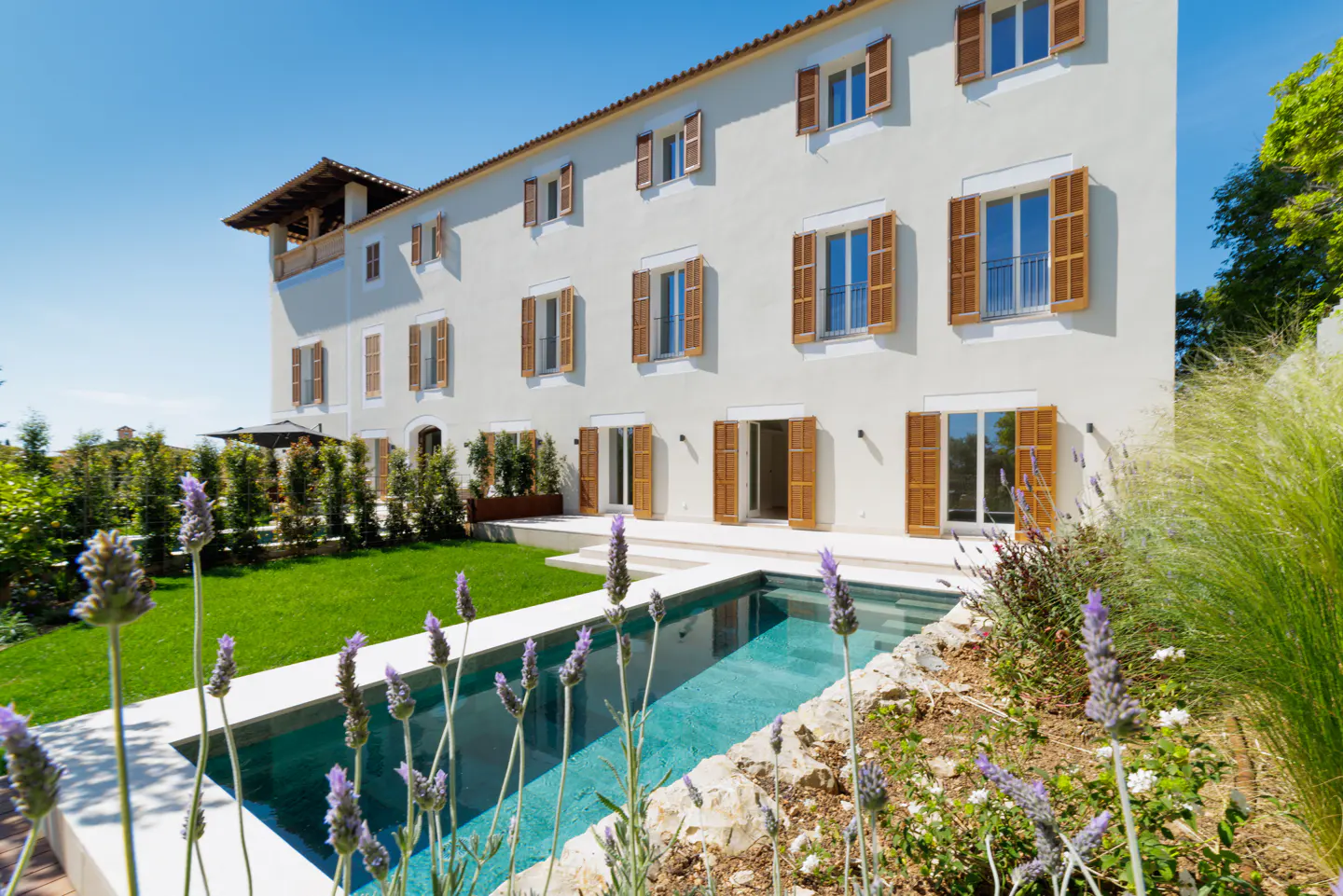 Exterior view of a large, white house with brown shutters, a pool, and lavender flowers in the foreground.