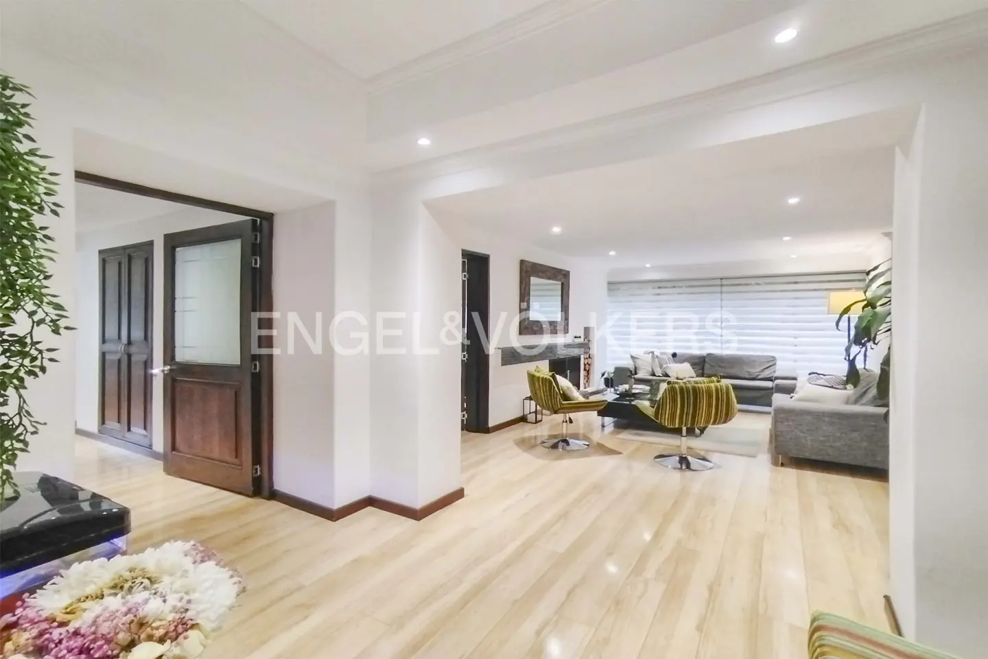 A bright, open-concept living space with light wood floors, white walls, and modern furniture. A dark wood door is on the left.