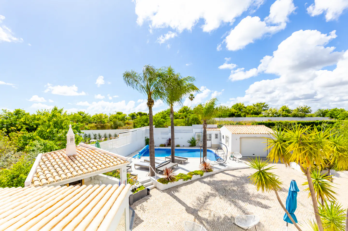 Aerial view of a backyard with a pool, palm trees, lounge chairs, and a white tiled patio under a blue sky with white clouds.