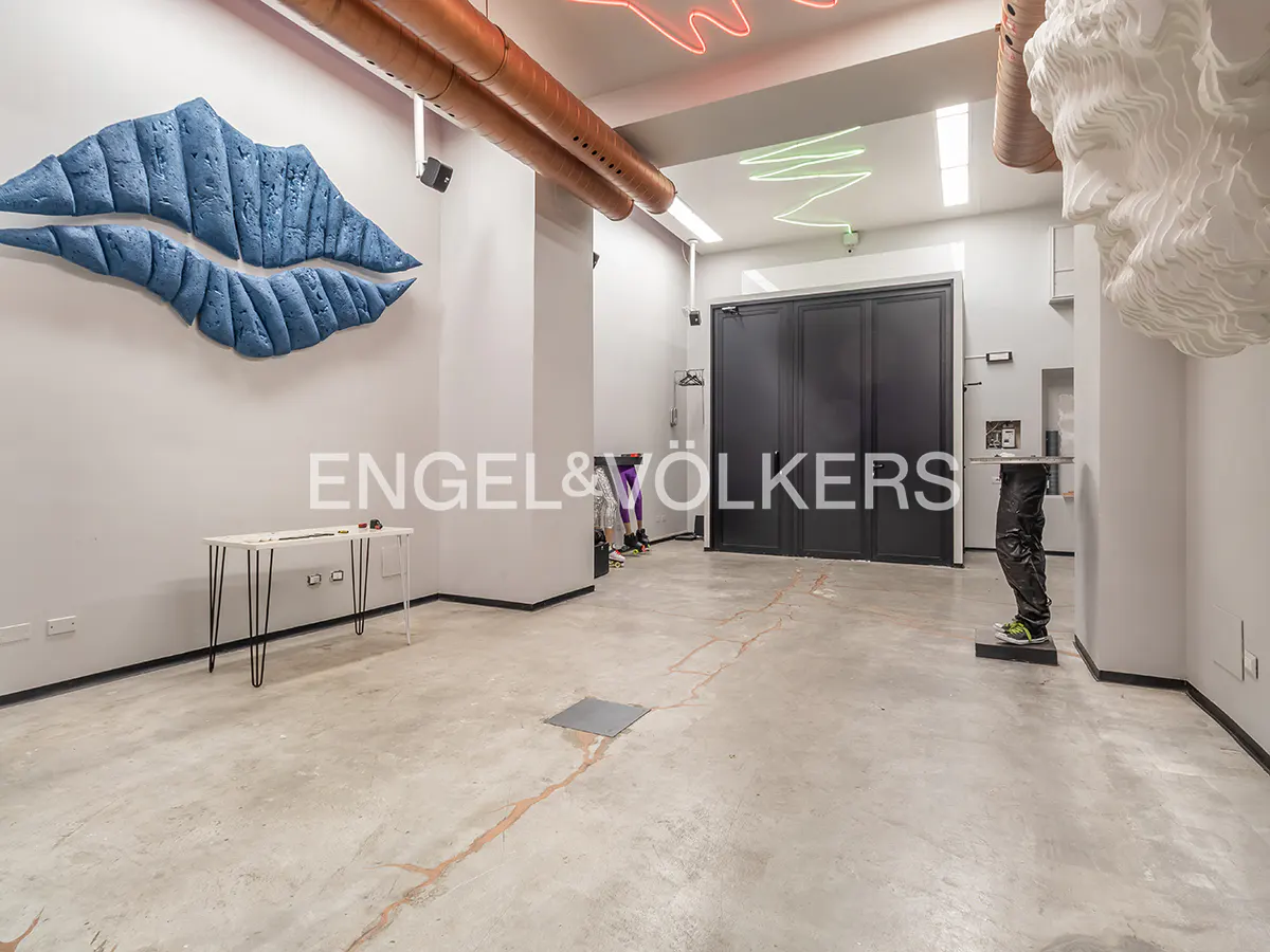 Bright, modern space with concrete floors, white walls, and art. A blue lip sculpture hangs above a white table. Neon lights adorn the ceiling.