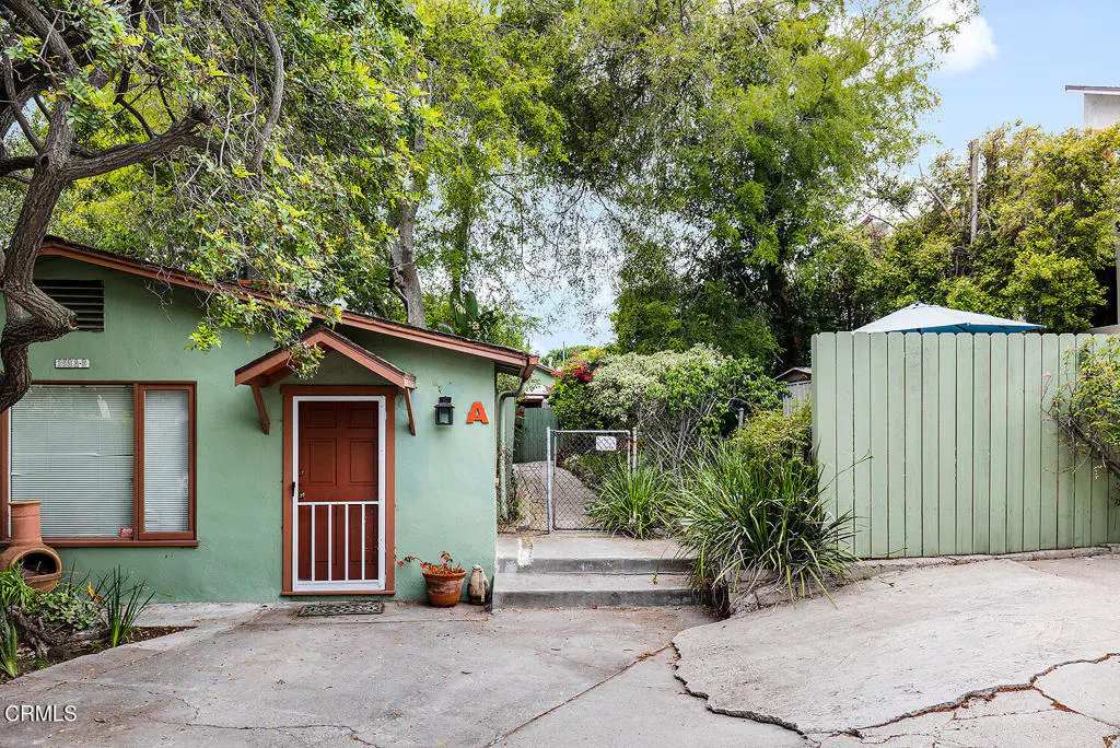 A quaint, light green house with a brown door and trim, surrounded by lush greenery and a light green fence.