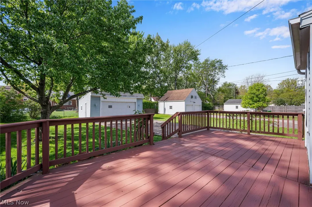 A brown wooden deck with a railing overlooks a green lawn and two white garages under a blue sky.