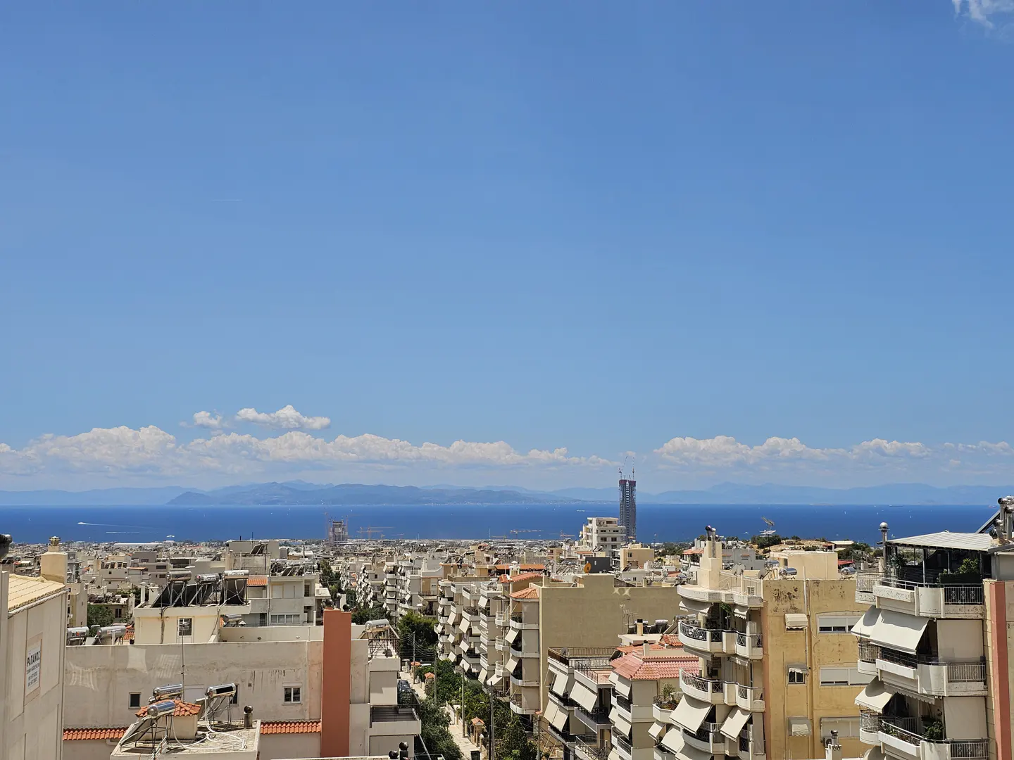 Elevated view of a dense Mediterranean city with beige buildings, balconies, and a distant blue sea under a clear sky.