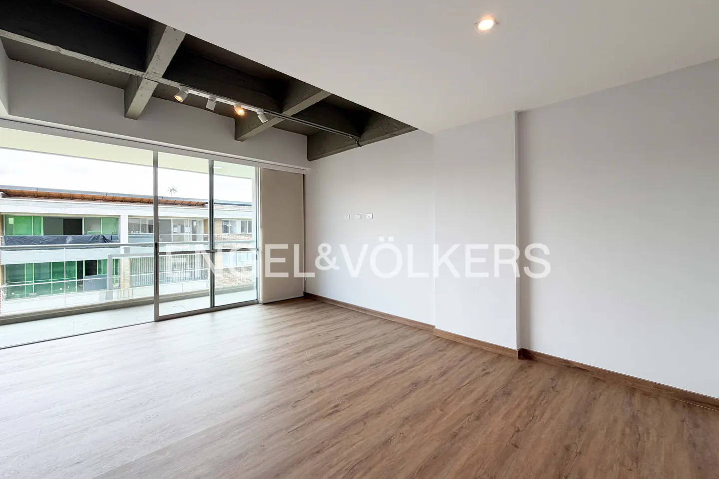 Empty room with wood floors, white walls, and a large sliding glass door to a balcony. Gray ceiling beams with track lighting. Engel & Völkers logo.