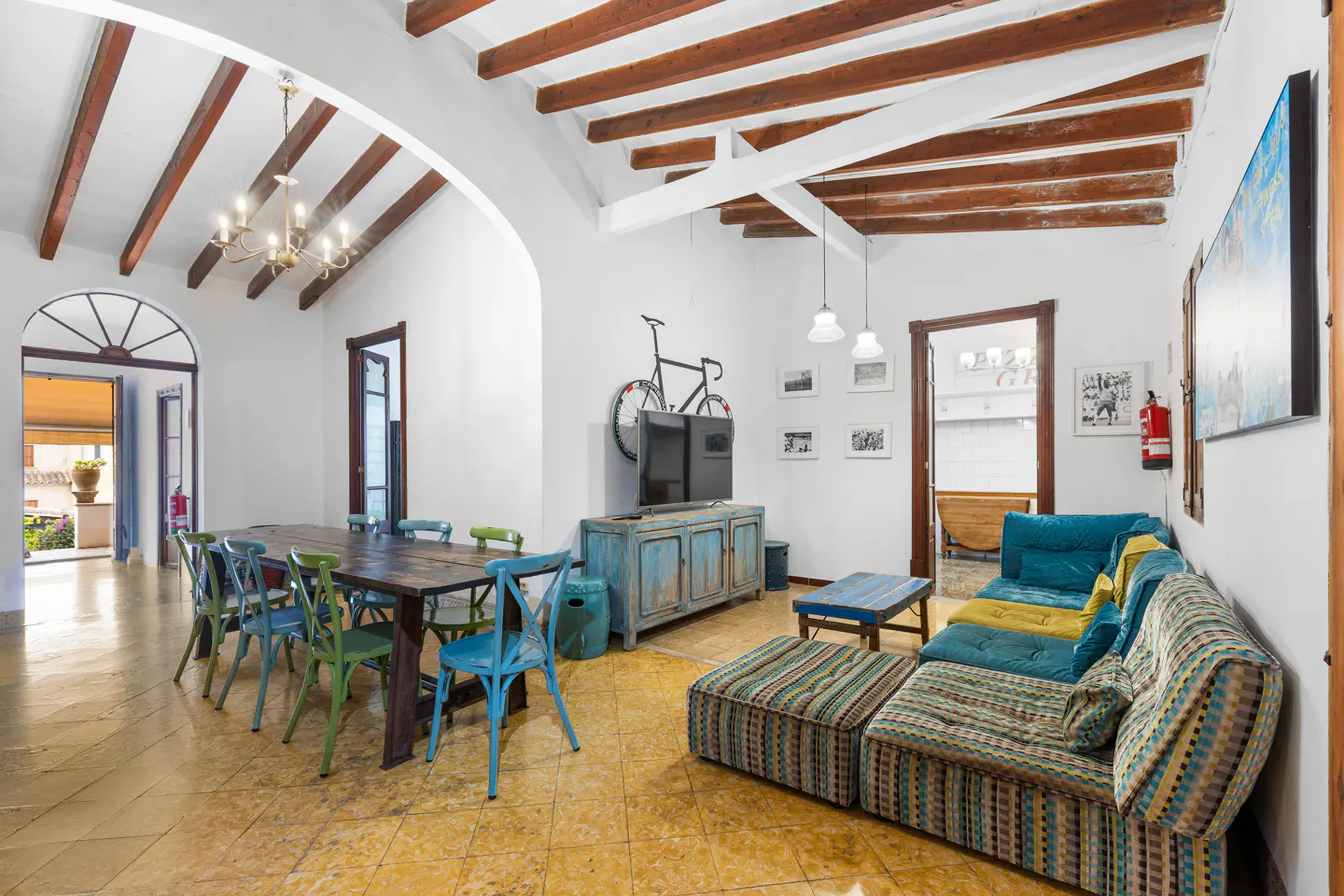 Bright living room with a wooden table, colorful chairs, and a patterned sofa. A bicycle hangs above a blue cabinet with a TV. Beamed ceiling.