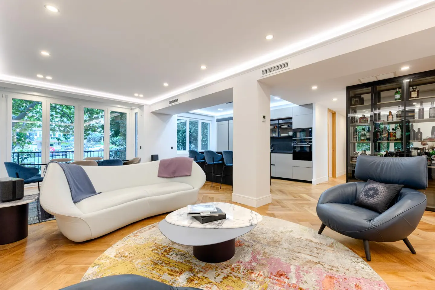 Bright, modern living room with white sofa, blue chair, and colorful rug. Kitchen and bar area visible in the background.