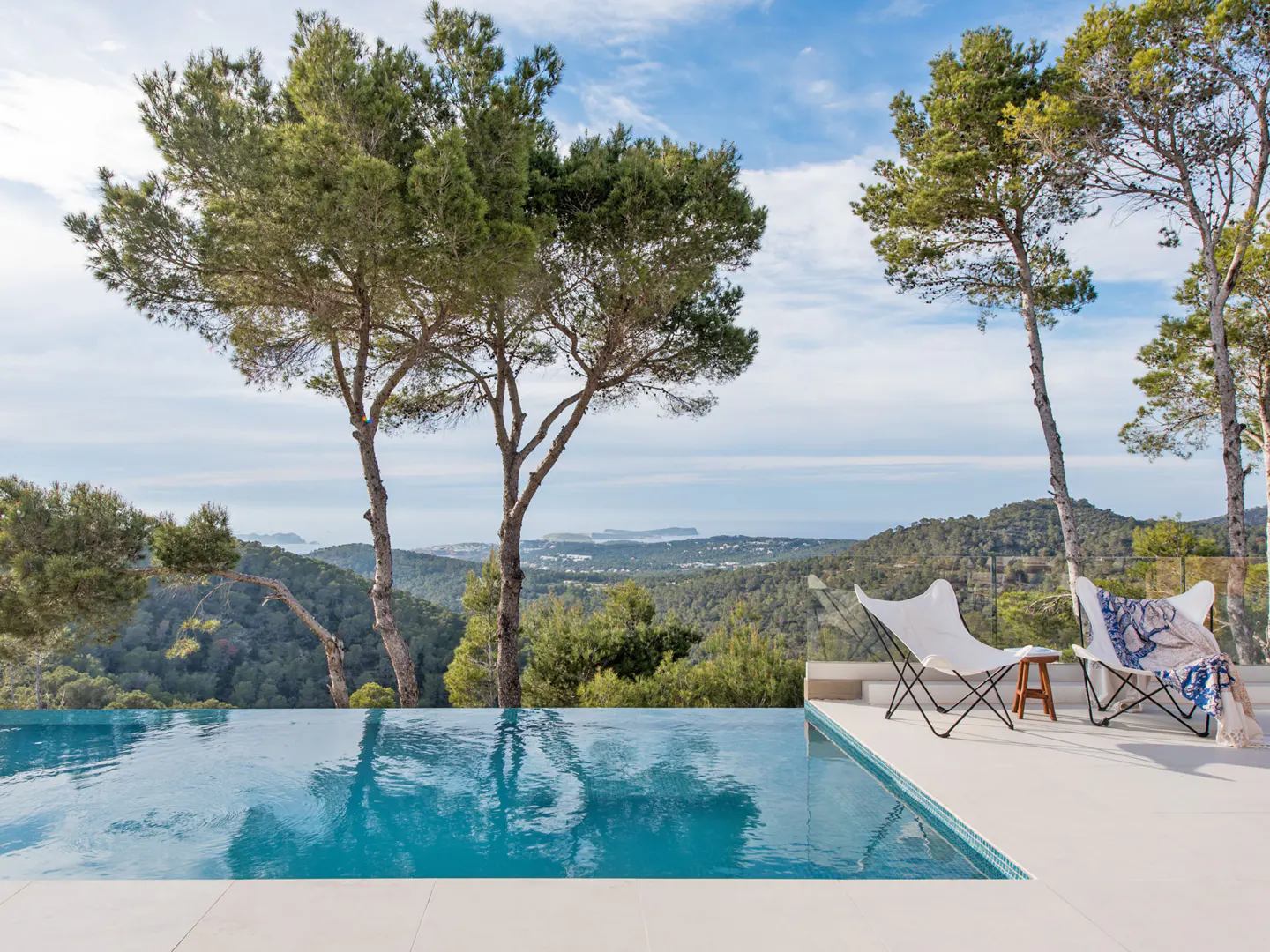 Infinity pool with turquoise water overlooks green hills and a distant island under a blue sky. Two white butterfly chairs sit on the patio.