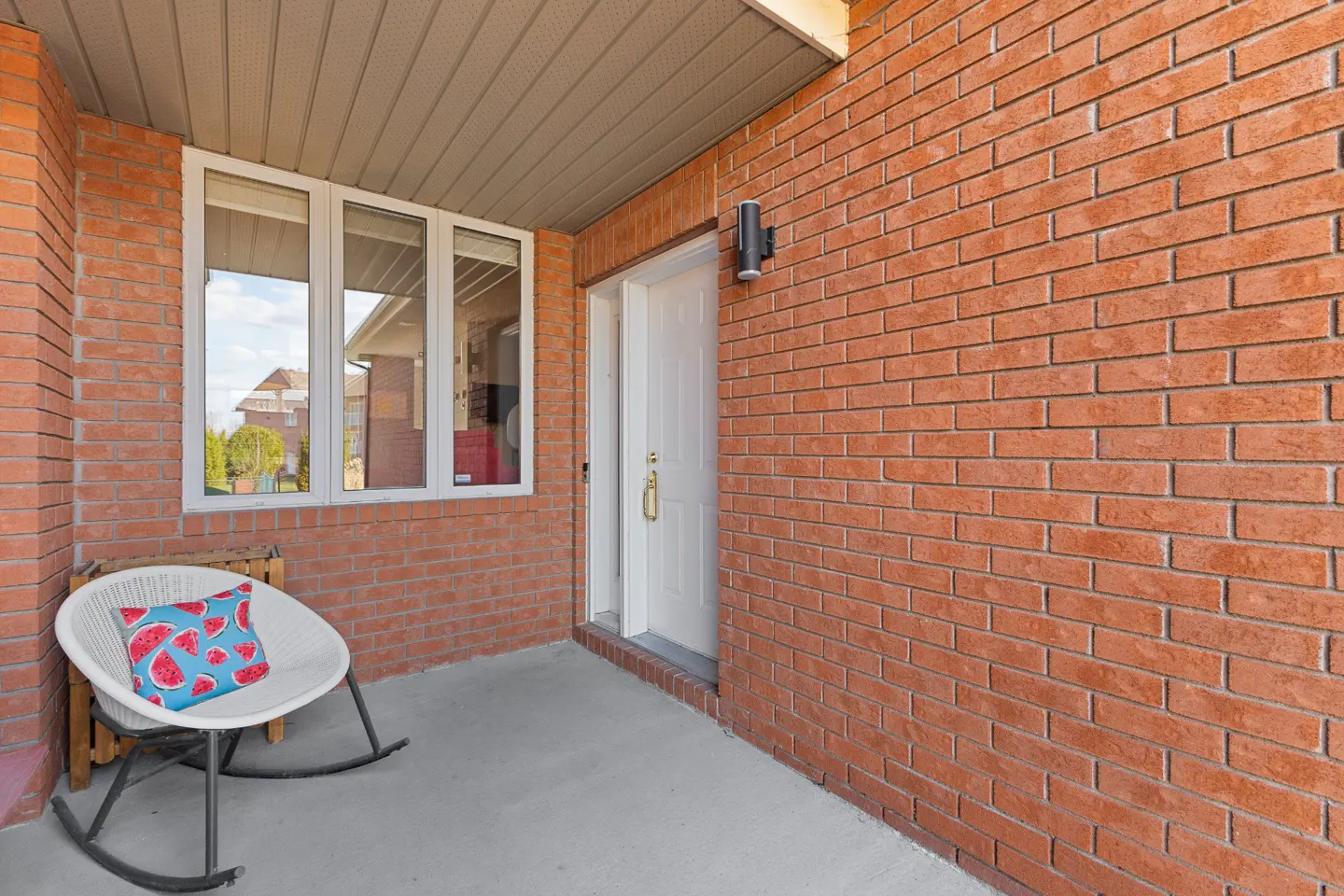 Covered porch with red brick walls, a white front door, and a white rocking chair with a watermelon pillow.