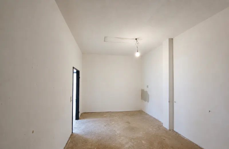 Empty room with white walls, concrete floor, a single hanging light bulb, and a doorway leading to another area.