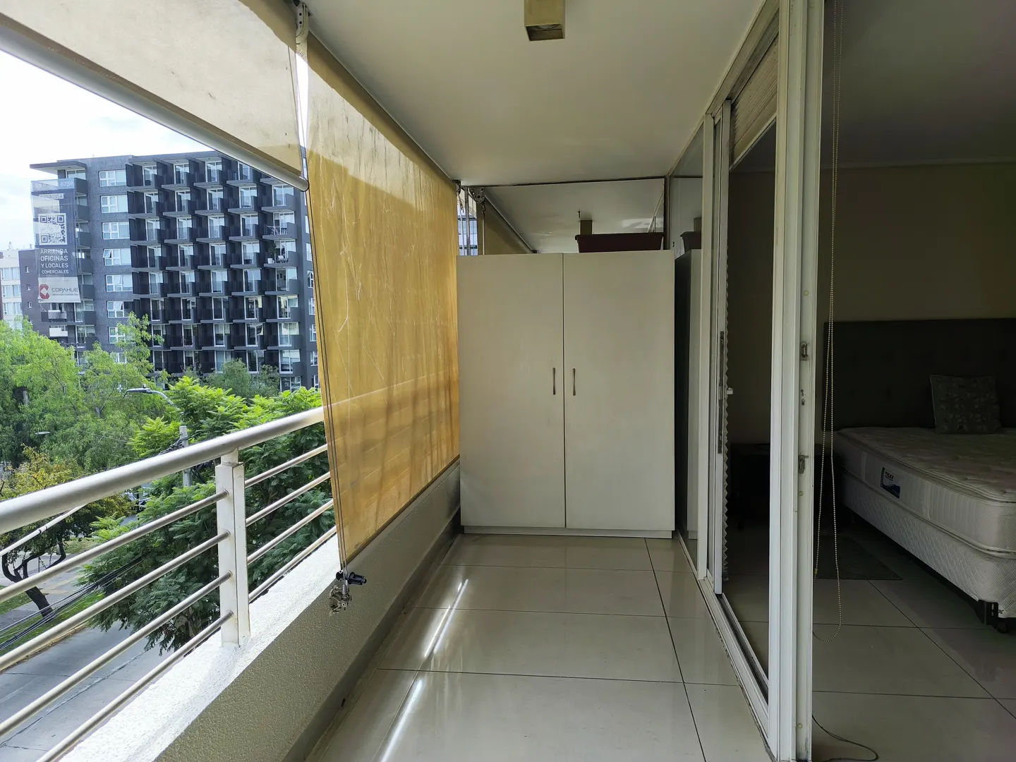 A balcony with a view of a city building and trees, featuring a white wardrobe and sliding glass doors.