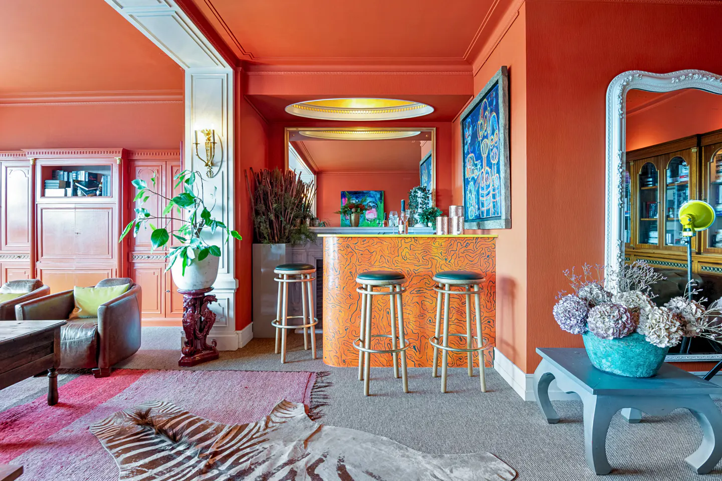 Interior view of an orange room with a bar, stools, zebra rug, and seating area. A large mirror reflects the room.