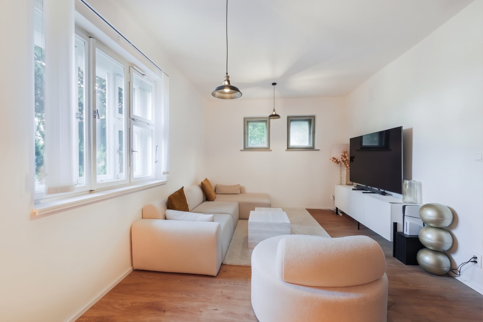 Bright living room with a beige sectional sofa, white coffee table, large TV on a white console, and modern pendant lights. [AI-generated text]