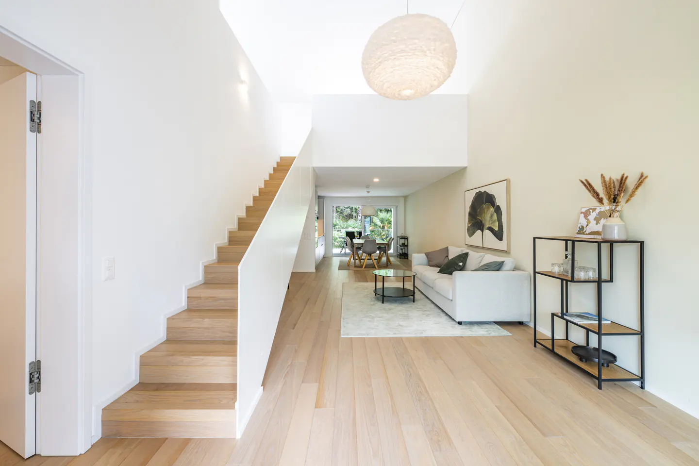 Bright, modern living room with light wood floors, white walls, and a staircase. A white sofa and black shelving unit add contrast.
