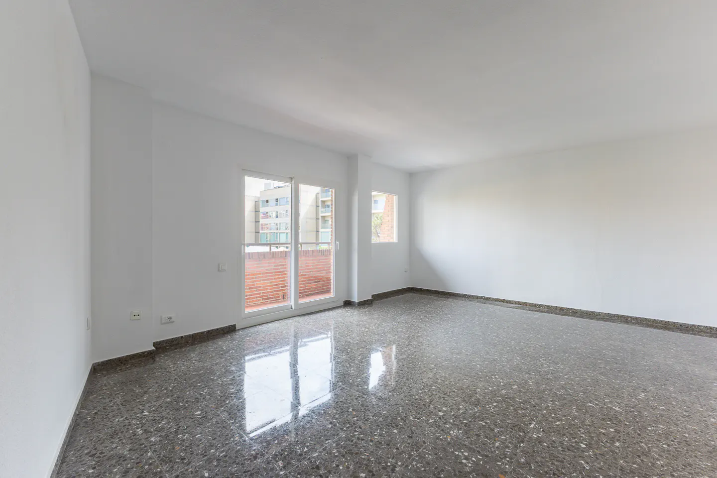 Empty room with white walls, polished grey speckled stone flooring, and a glass door leading to a brick balcony.