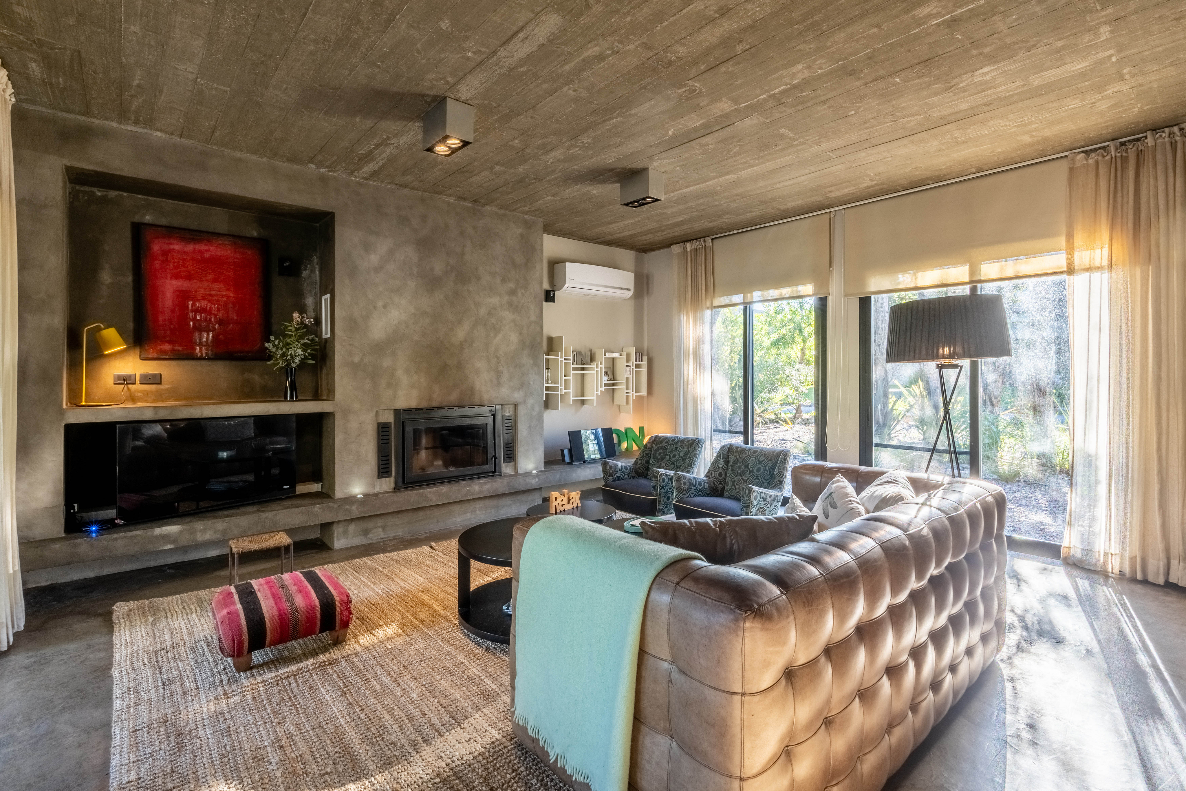 Living room with concrete walls, a brown leather sofa, a fireplace, and large windows with sheer curtains.