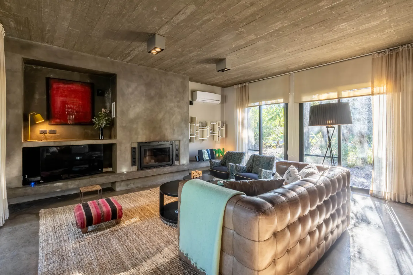 Living room with concrete walls, a brown leather sofa, a fireplace, and large windows with sheer curtains.