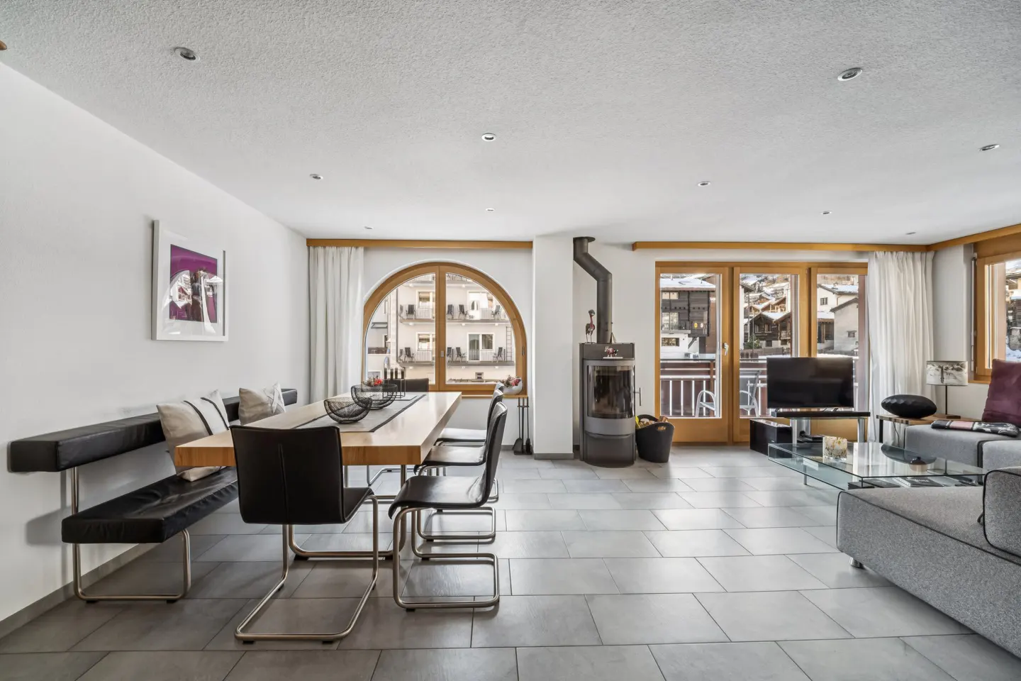 Bright living room with gray tile floor, wood dining table, black chairs, and a fireplace. Large windows offer a view of buildings.