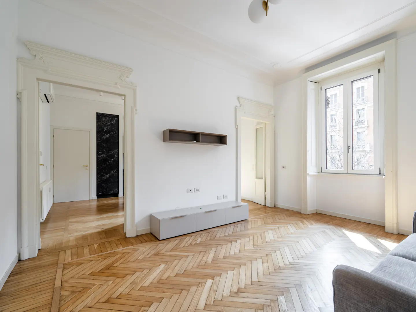 Bright, empty living room with herringbone wood floors, white walls, and large window. Gray media console and floating shelf. Doorways with decorative molding.
