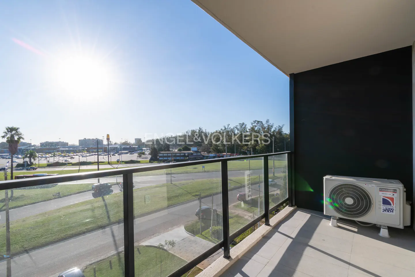Balcony view with glass railing overlooking a road, buildings, and blue sky. An air conditioner sits on the balcony floor.