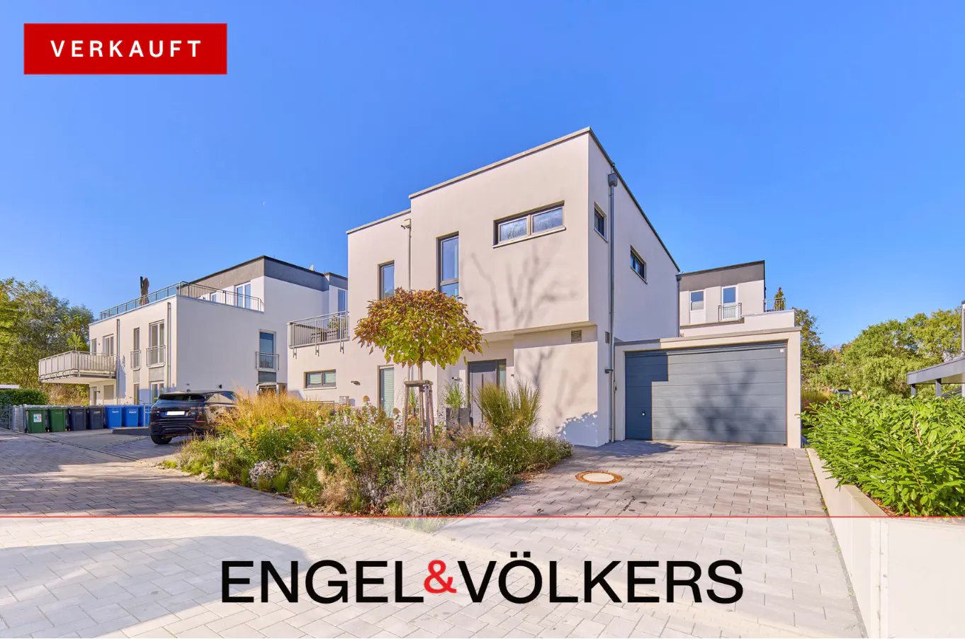 Modern two-story white house with a gray garage door and a small tree in front. "VERKAUFT" banner in the upper left corner.