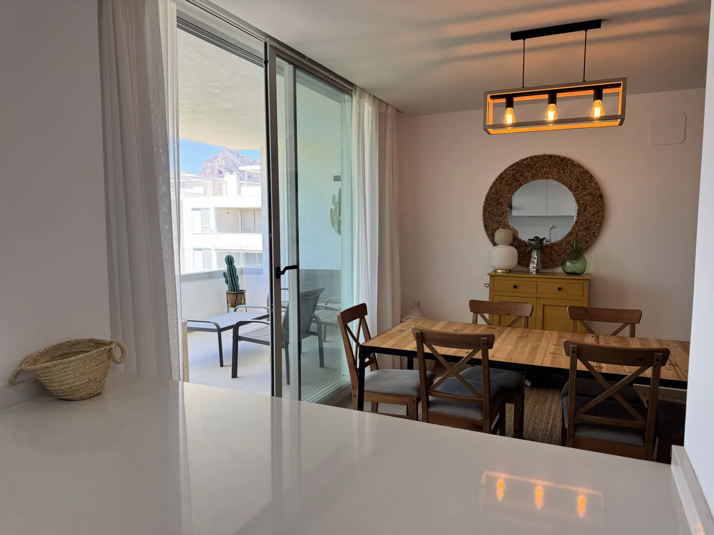 Bright dining room with a wooden table, chairs, and a yellow cabinet under a round mirror. Balcony with mountain view visible through sliding glass doors.