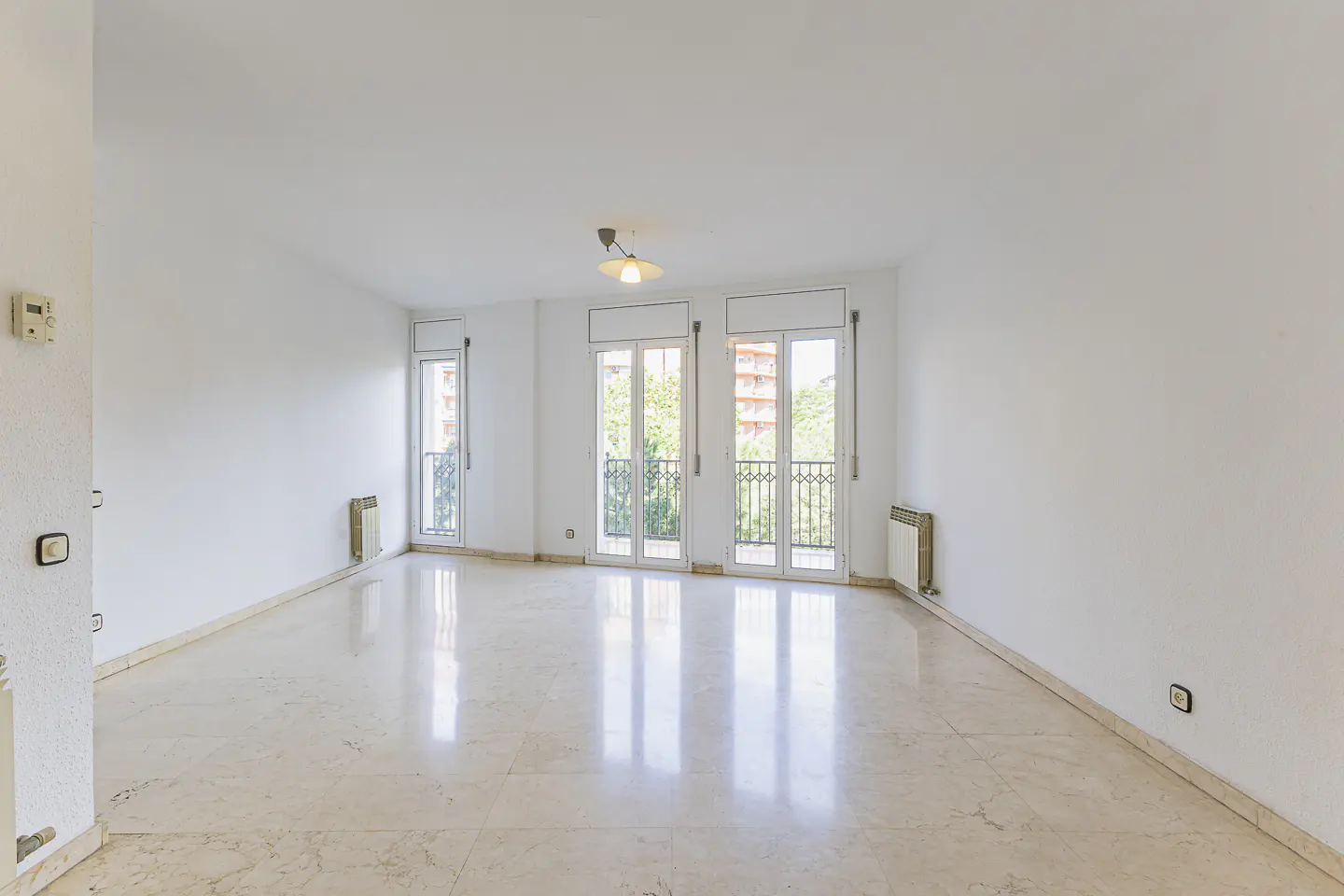 Bright, empty room with white walls, marble floor, and three sets of doors leading to balconies. Radiators are under the windows.
