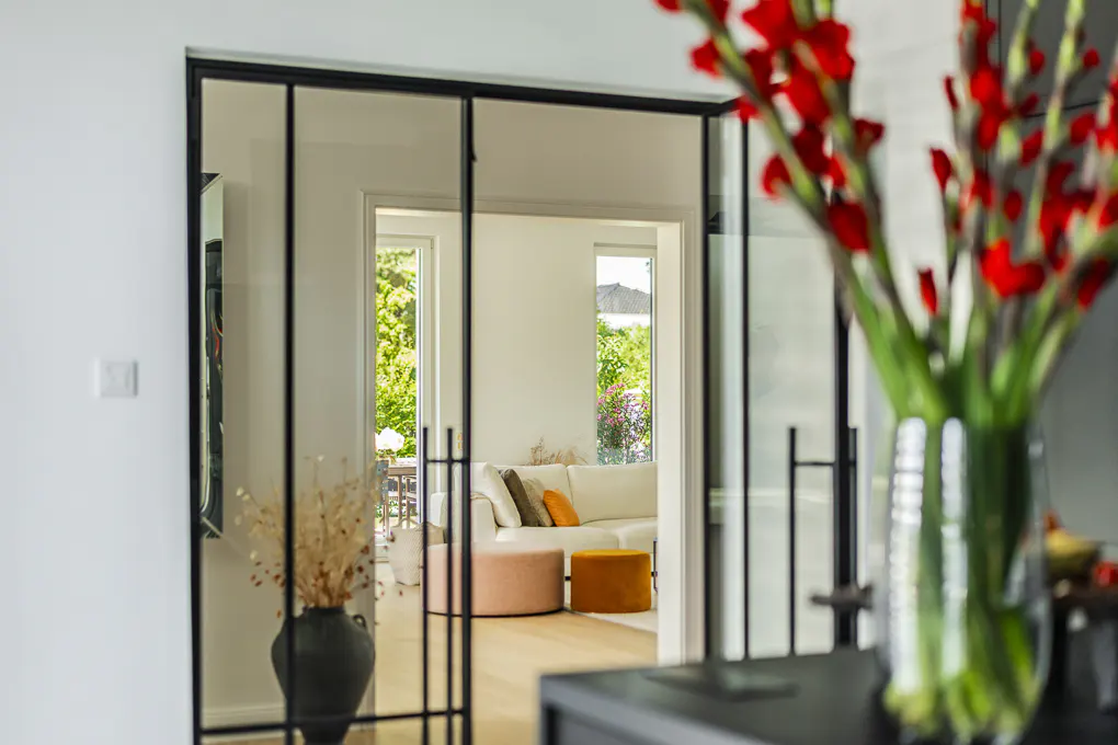 Interior view through black-framed glass doors into a bright living room with a white sofa, colorful pillows, and outdoor greenery. Red flowers in a vase are in the foreground.