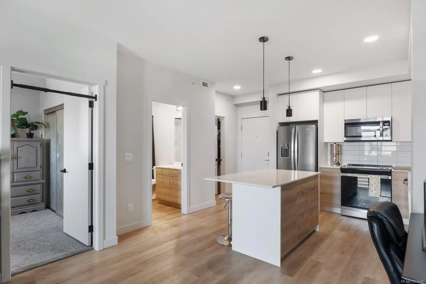 Bright, modern condo interior with wood floors, white walls, and an open kitchen with island. Bedroom and bathroom visible through doorways.