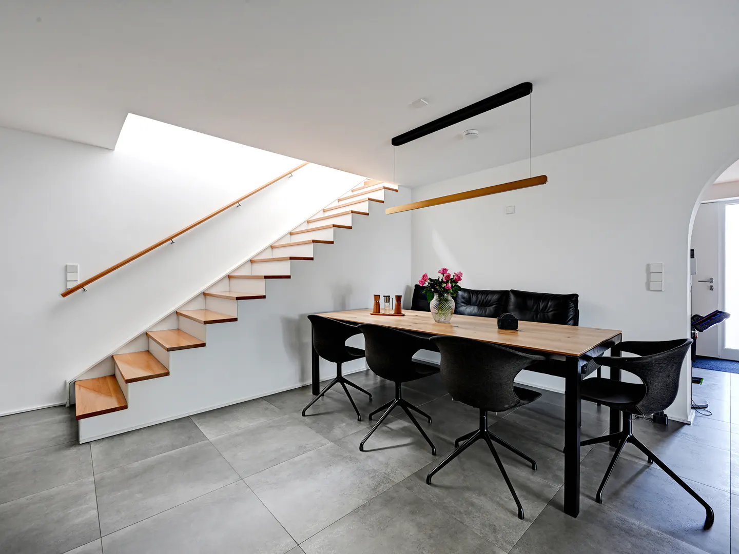 A modern dining area with a wooden table, black chairs, and a black leather sofa against a white wall. A staircase with a wooden handrail is visible.