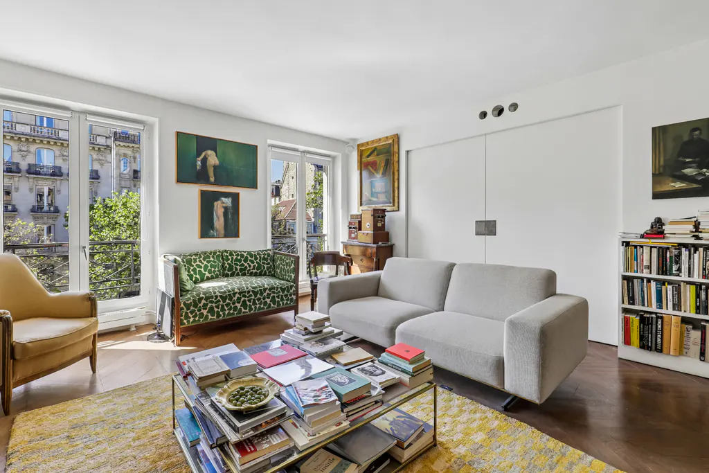 Bright living room with a grey sofa, green patterned loveseat, and a glass coffee table piled with books. Windows show a city view.