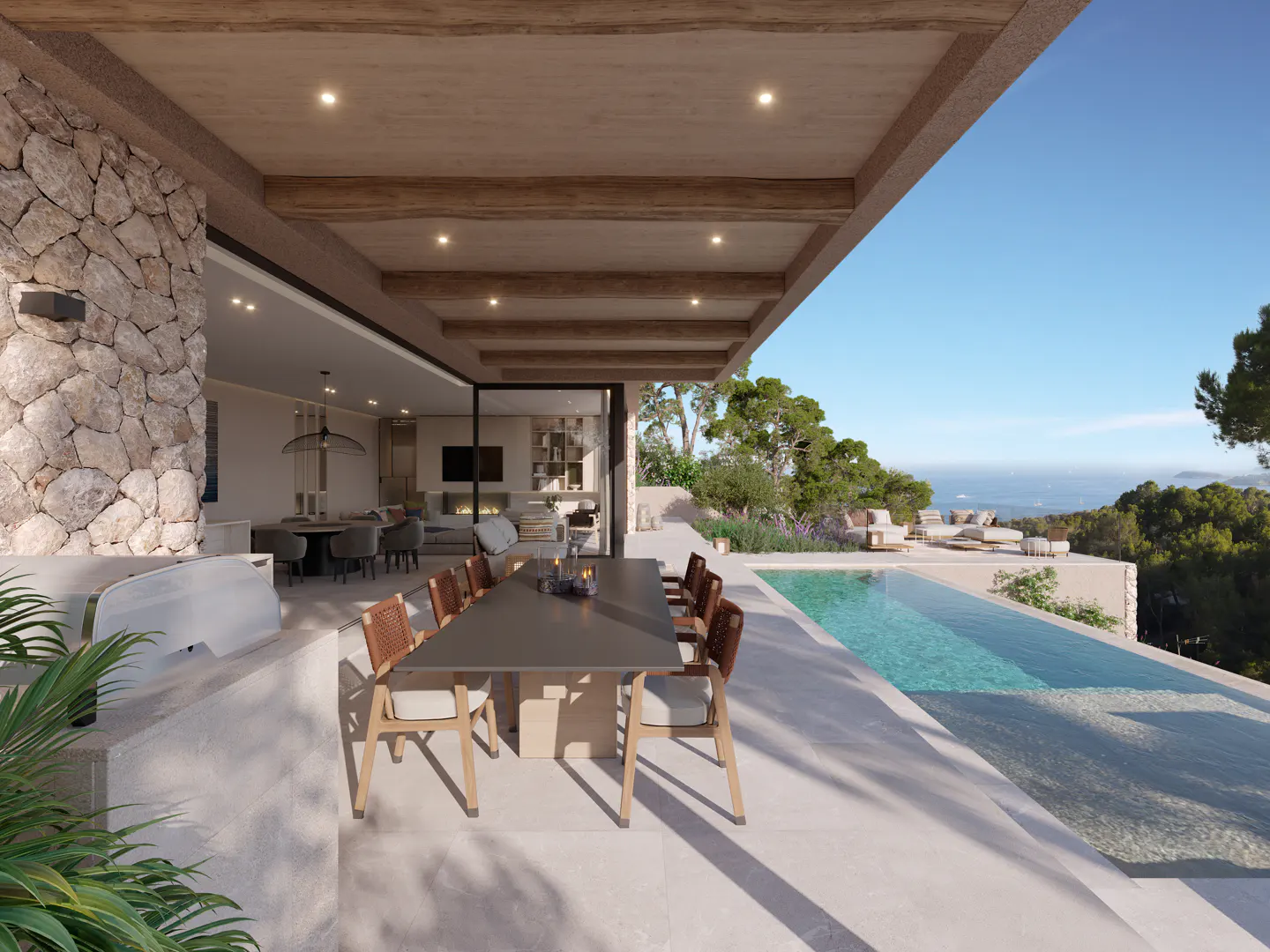 Outdoor dining area with a long table and chairs next to a pool overlooking the ocean. The house has a stone wall and wooden beams.