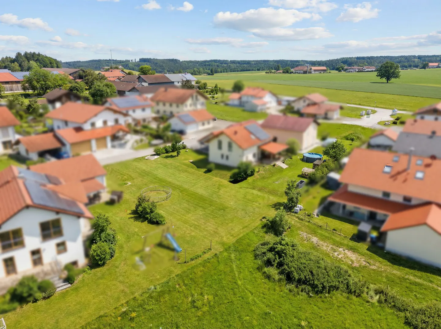 Aerial view of a quaint village with houses with red roofs, green lawns, and a field in the background under a blue sky.