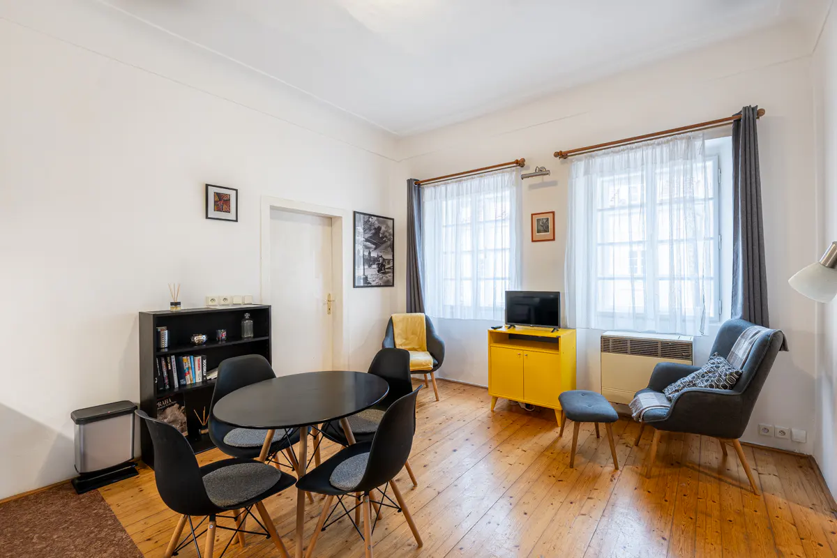 Bright living room with wood floors, black table and chairs, yellow cabinet, and two windows with white curtains.