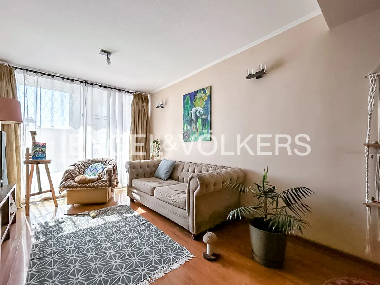 Living room with a beige sofa, patterned rug, armchair, and a painting. Natural light filters through sheer curtains.