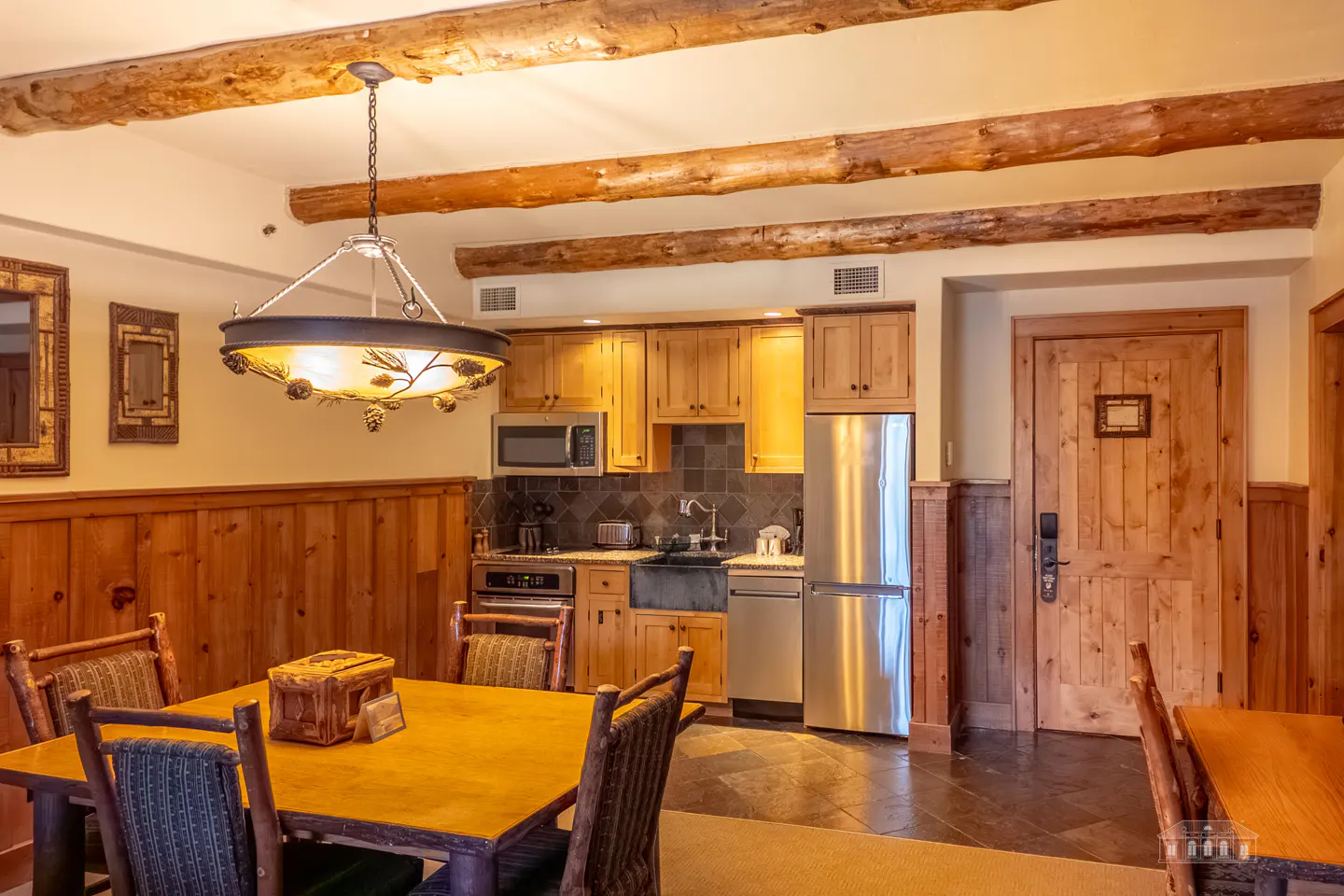 Rustic kitchen and dining area with wood paneling, beams, and a pinecone chandelier. Stainless steel appliances and a wooden dining table.