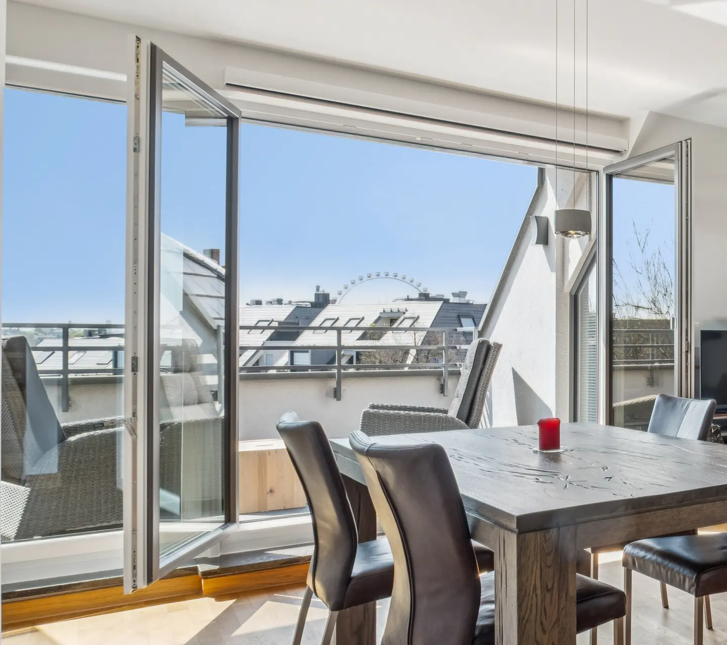 Bright dining room with open doors to a balcony. A dark wood table and chairs sit in front of the open doors, with a view of rooftops and a Ferris wheel.