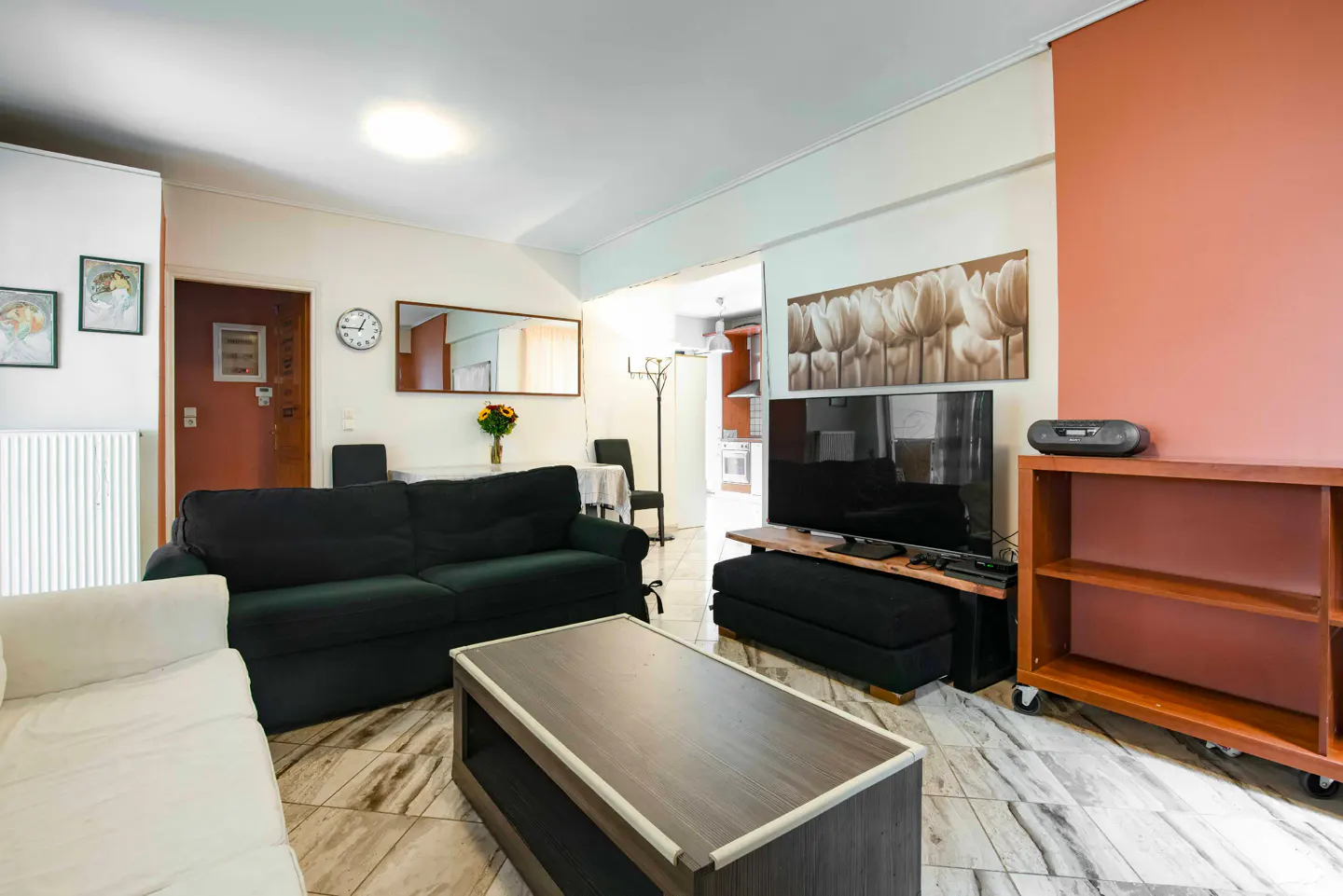 A living room with a black sofa, a dark wood coffee table, and a TV on a wooden stand. A bookshelf is on the right.