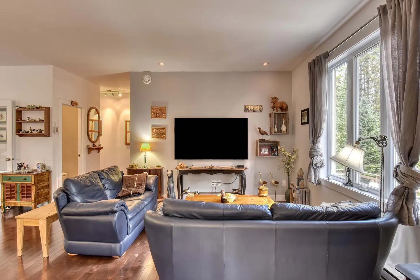 Living room with two blue leather sofas, wood floors, and a large window with gray curtains. A TV hangs on the wall.