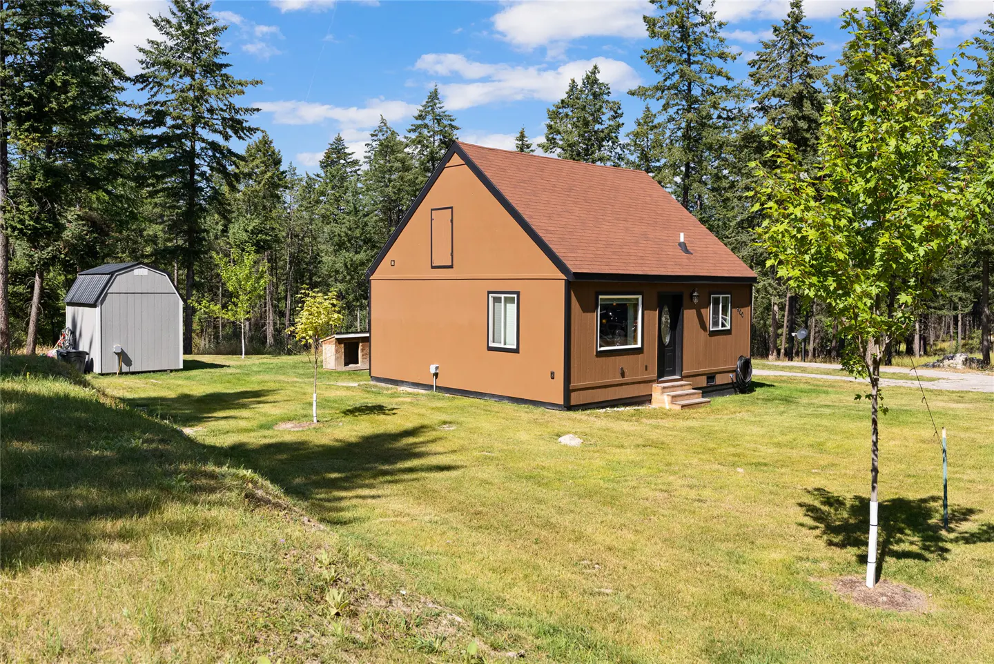 A brown, one-story house with a brown roof sits on a green lawn with trees in the background. A gray shed is on the left.