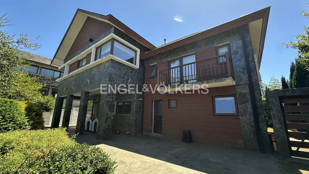 Two-story house with stone and wood facade, balcony, and large windows under a blue sky. Engel & Völkers logo visible.