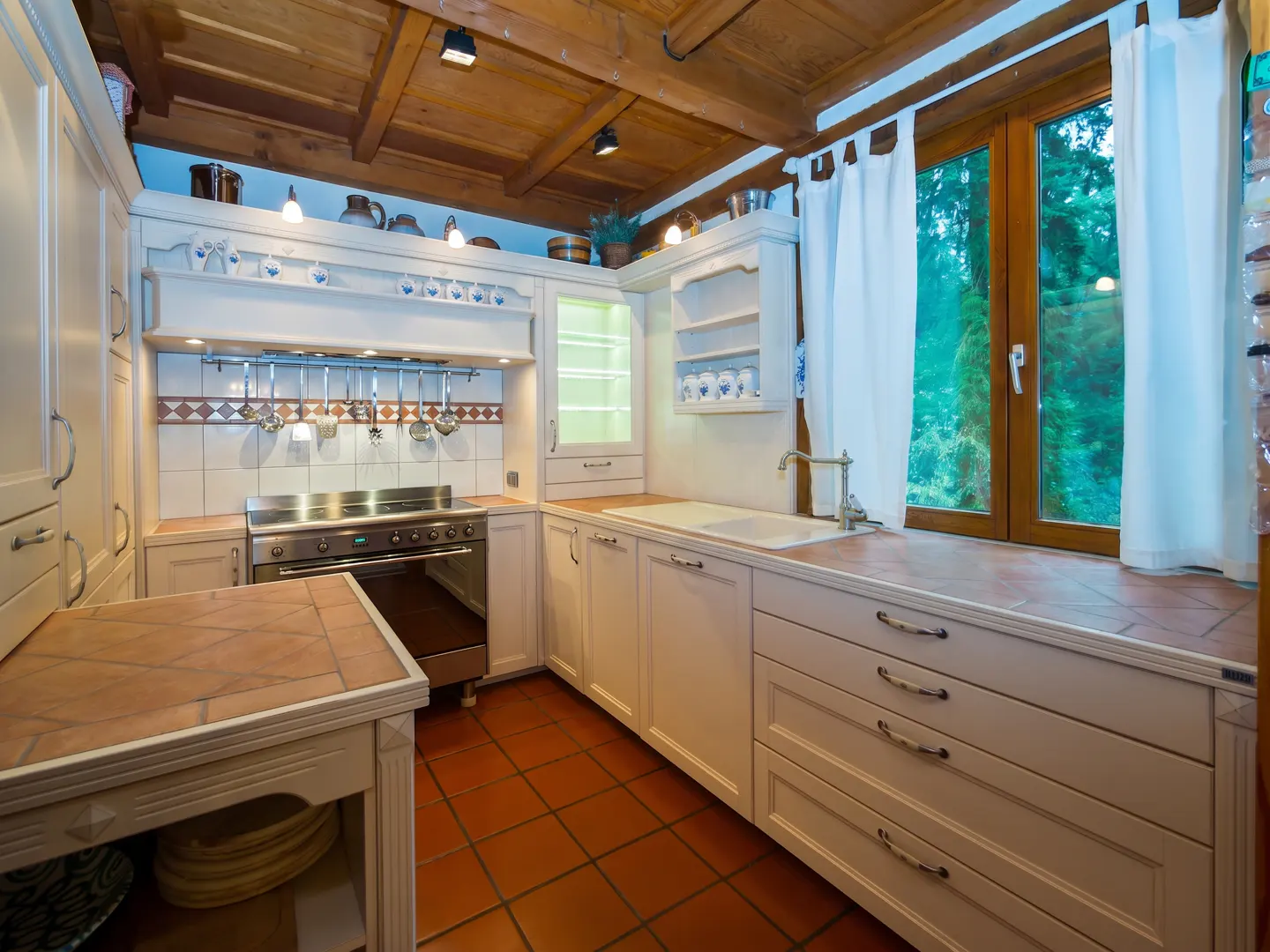 A bright kitchen with white cabinets, a stainless steel stove, and terracotta tile floors. A window with white curtains overlooks a green forest.