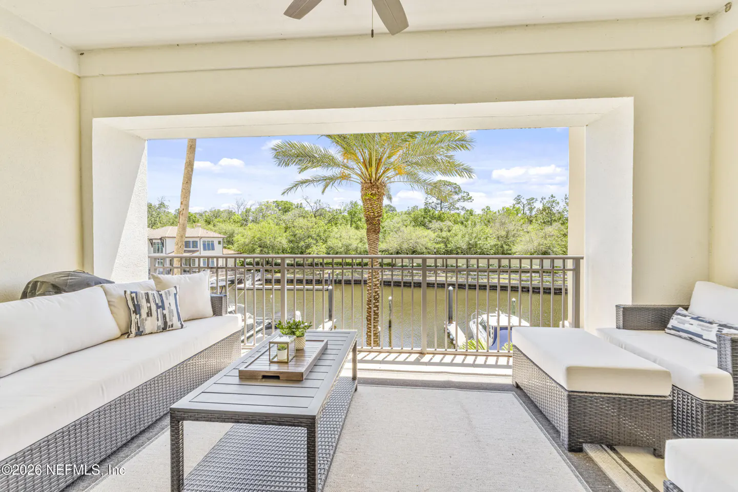 A waterfront balcony with wicker furniture, white cushions, and a palm tree view.