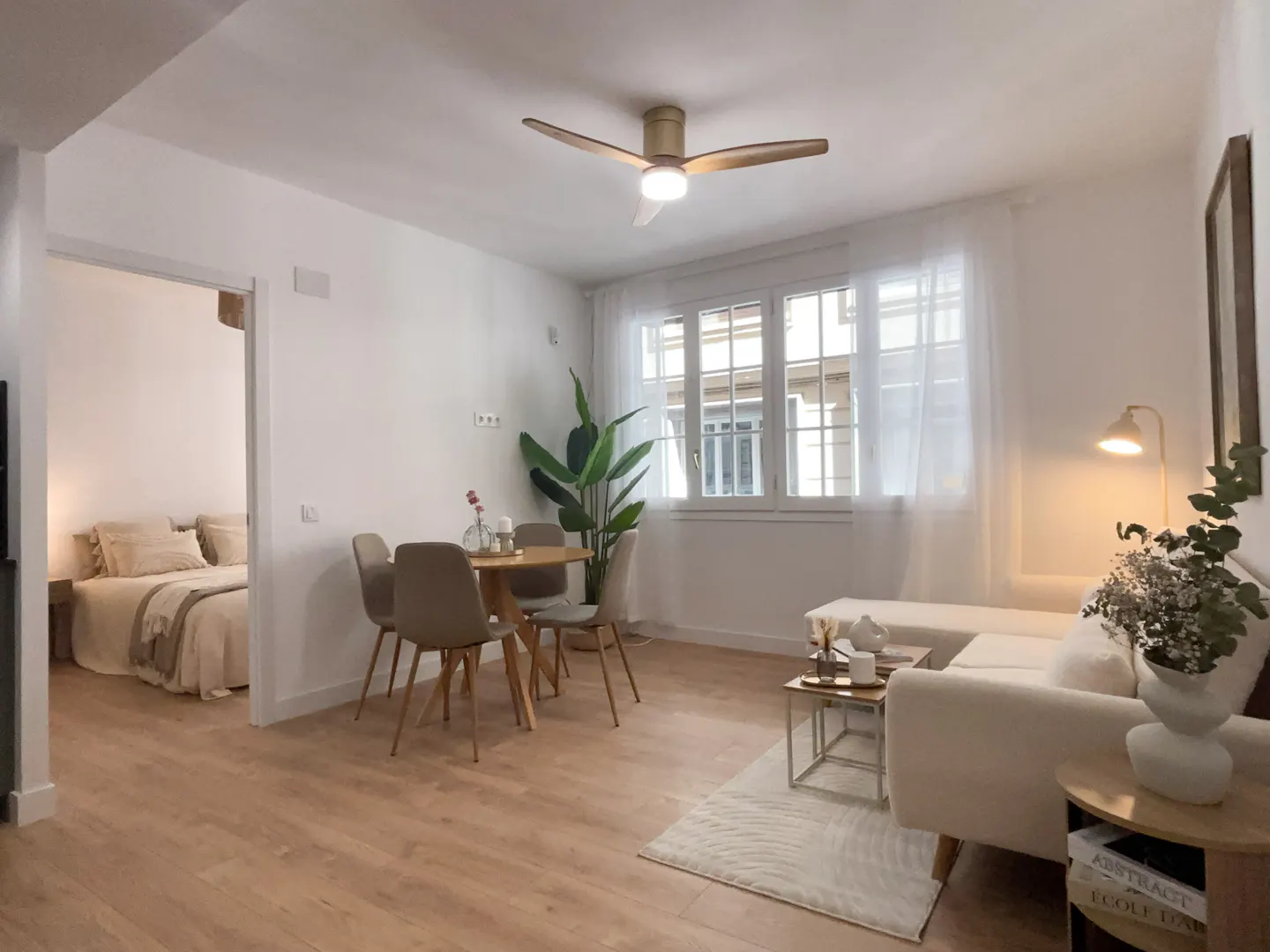 Bright, modern apartment interior with wood floors, white walls, and a ceiling fan. A dining table, sofa, and bedroom are visible.